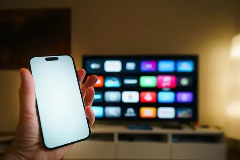 Celular em primeiro plano diante de televisão com aplicativos ao fundo.